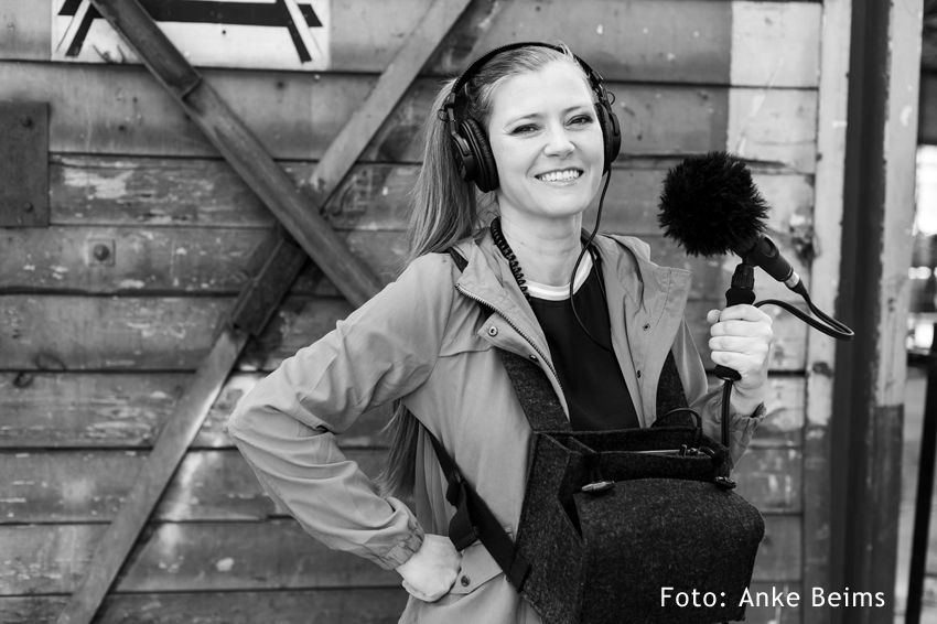 Foto: schwarz-weiß Porträt Susanne Franzmeyer mit Mikrofon und Kopfhörern vor Bretterwand 